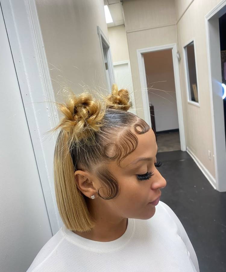 Mulher loira com cabelo liso e baby hairs estilizados. Ela usa maquiagem brilhante, cílios longos, piercing no nariz, colar dourado e camiseta preta. A luz do sol ilumina seu rosto enquanto está sentada dentro de um carro.





