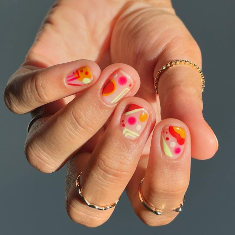 Mão com unhas curtas decoradas com desenhos abstratos em tons de laranja, rosa e amarelo sobre base transparente. A pele está iluminada pela luz solar, destacando anéis dourados delicados. Fundo cinza desfocado, criando um contraste suave.