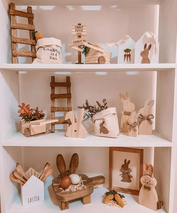 Prateleiras com decoração temática de Páscoa, incluindo coelhos de madeira, sacos rústicos, miniaturas de cenouras, escadas decorativas, arranjos florais, moldura com coelho, carrinho de mão e ovos em plataforma de madeira. Combinação rústica e acolhedora em tons naturais.