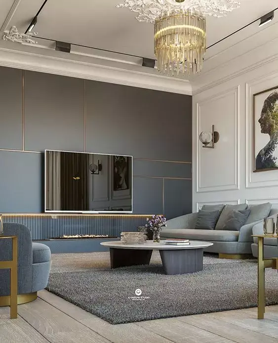 Sala de TV com sofás, mesa e tapete cinzas, lustre, parede escura com boiserie dourado e parede branca com moldura de mesma cor 