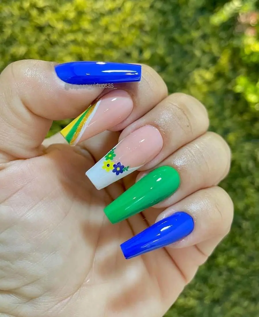 Imagem de mão com unhas decoradas para a Copa do Mundo 2022. Cores usadas, verde, amarelo, azul, branco e glitter dourado. Polegar: azul. Indicador: branco com glitter verde, dourado e azul. Dedo Médio, verde. Anelar: branco com glitter verde, dourado e azul. Mínimo: azul.