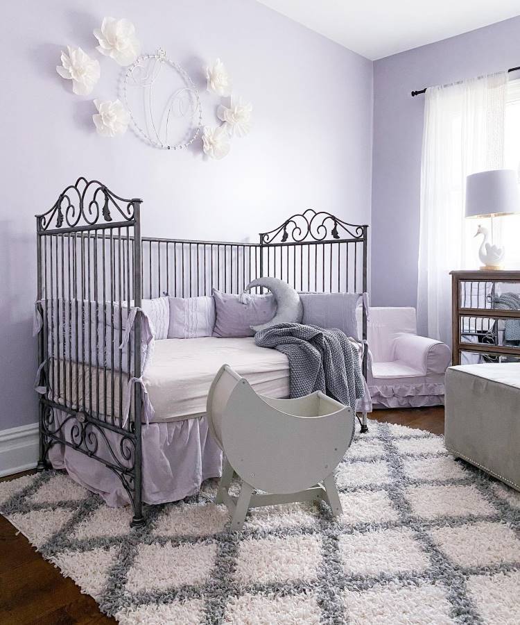 A imagem apresenta um quarto infantil em tons de lavanda, com um berço de ferro decorativo, almofadas suaves e detalhes delicados como flores na parede. Dicas de decoração para quem tem TDAH incluem cores claras e organização visual simples.