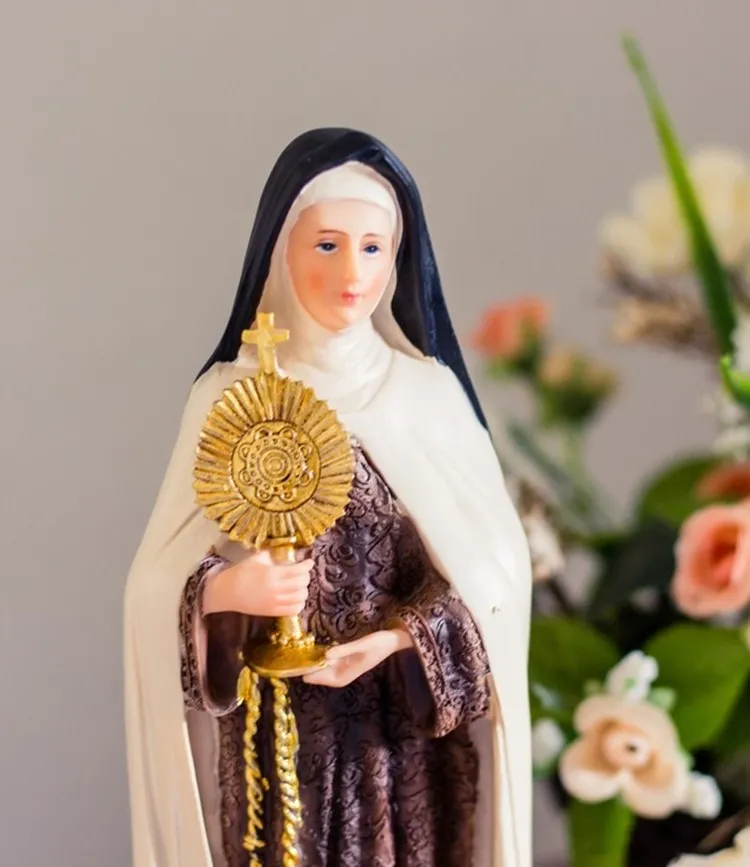 A estátua de Santa Clara de Assis está vestida com um hábito marrom e véu preto. Ela segura um ostensório dourado com uma cruz no topo. O fundo é cinza claro, e há flores coloridas ao redor, incluindo rosas e lírios, destacando a serenidade da santa.


