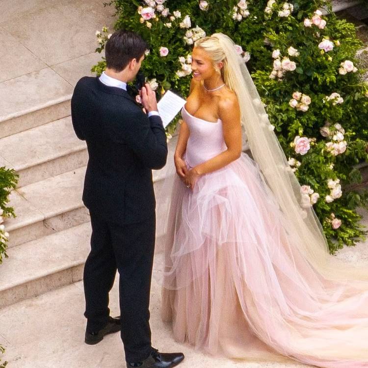 Uma noiva loira, sorrindo e usando um vestido de noiva rosa tomara que caia com saia de tule esvoaçante e véu, olha para o noivo de terno escuro que segura um papel. Um arbusto florido está ao fundo.







