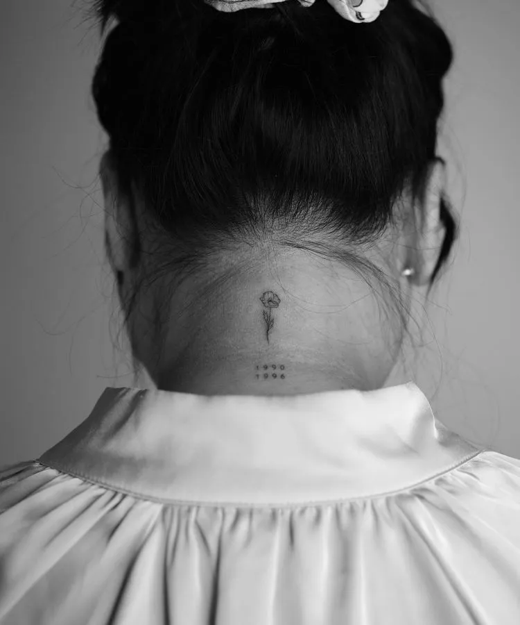 A imagem mostra uma pessoa de costas com cabelo preso em um coque. Ela tem uma tatuagem no pescoço de uma pequena flor delicada com um caule fino e duas datas abaixo dela. A tatuagem está localizada na base do pescoço, centralizada.
