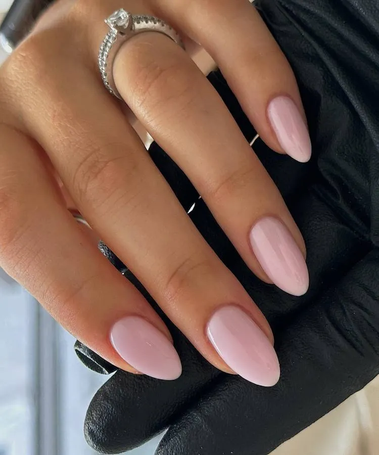 Unhas médias a grandes, no formato amendoada, na cor rosa claro. Uma mão com luva preta, possivelmente da manicure, segura esta mão esmaltada. A mão esmaltada possui um anel de brilhantes
