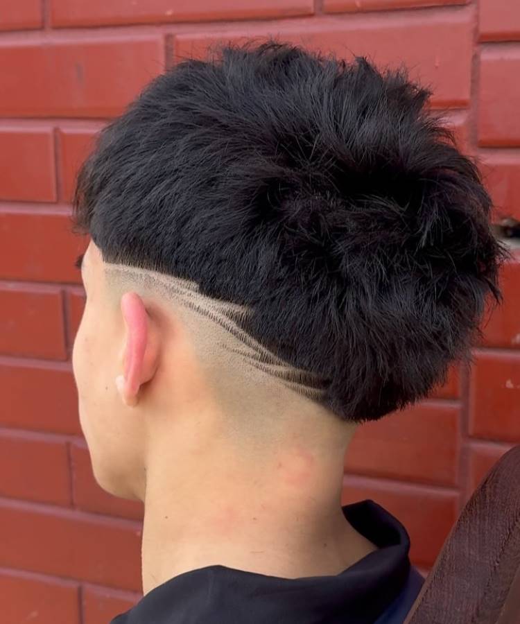 Um homem de costas com um **corte de cabelo bowl cut** e um undercut degradê. A parte de cima é escura, lisa e em forma de tigela, e as laterais e nuca têm um degradê (fade) com um design de linhas raspadas.