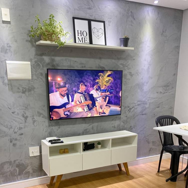 Sala pintada com cimento queimado, rack branco, TV, mesa branca com cadeira preta, quadros na decoração e vaso de Peperômia