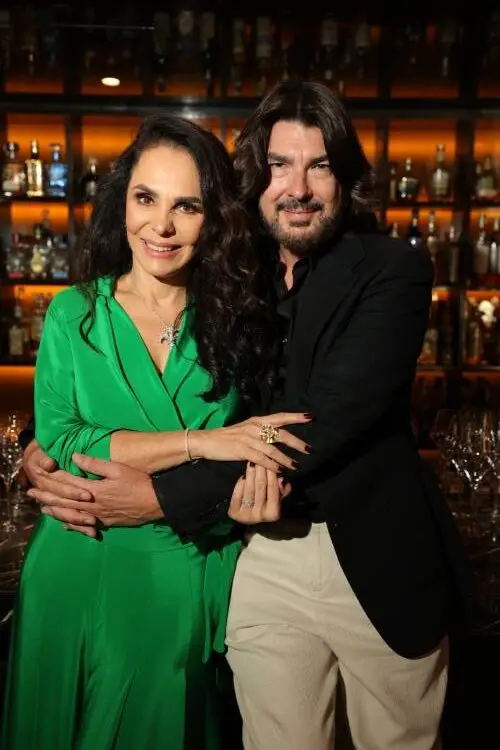 Cris Porto de verde e Stephane Rolland de terno preto e calça off-white abraçados em fundo de prateleira com bebidas