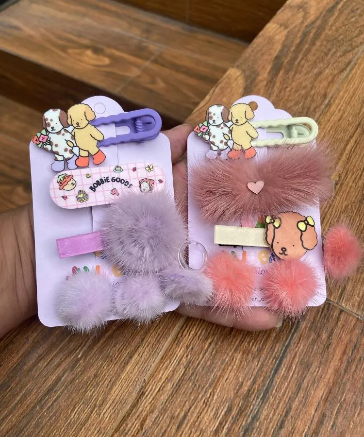 Dois pares de presilhas para cabelo em cartelas. Um par é lilás, com um cãozinho fofo e um pompom peludo. O outro par, rosa claro, também tem um cãozinho e pompons macios.

