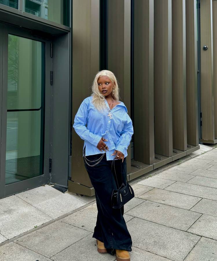 A imagem apresenta uma mulher vestindo uma calça jeans preta oversized, tendência 2025, com barra longa e cintura baixa, combinada com uma camisa azul listrada oversized. O look é complementado por botas caramelo, acessórios prateados e uma bolsa preta elegante.