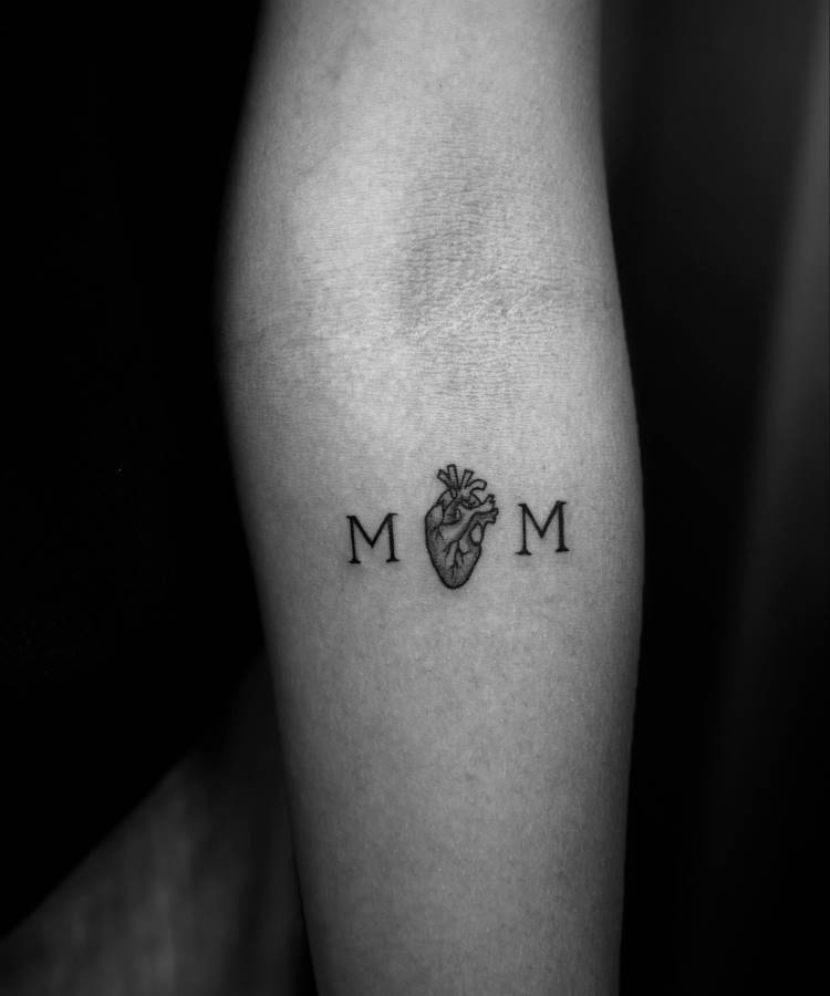 A imagem em preto e branco mostra uma tatuagem minimalista no antebraço de uma pessoa. A tatuagem consiste nas letras "M M" com um coração anatômico detalhado entre elas, formando a palavra "MOM". A pele ao redor é clara e lisa.







