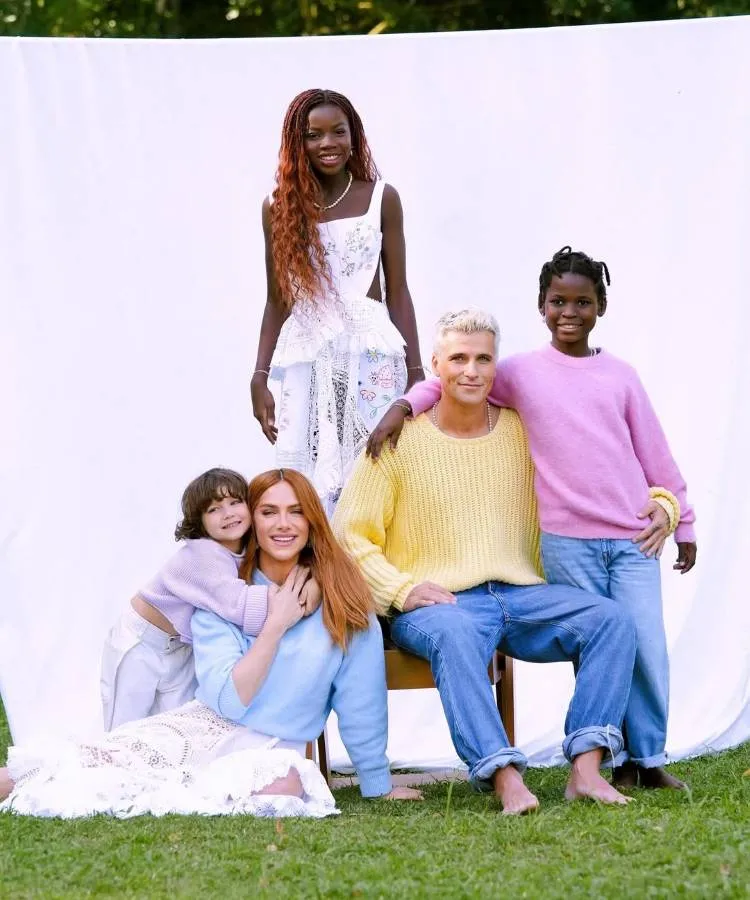 Uma família diversa posa contra um fundo branco. O pai loiro e a mãe ruiva estão sentados. Três crianças, uma com cabelo castanho e duas meninas negras (uma mais velha com tranças e vestido branco, outra com suéter rosa), os cercam. Nomes diferentes de filhos de famosos.

