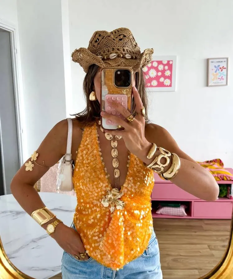 Mulher tira uma selfie no espelho, usando um chapéu de palha com búzios, um top de lantejoulas laranjas e jeans. Ela tem muitas joias douradas, incluindo um bracelete, um relógio e um colar com flores.