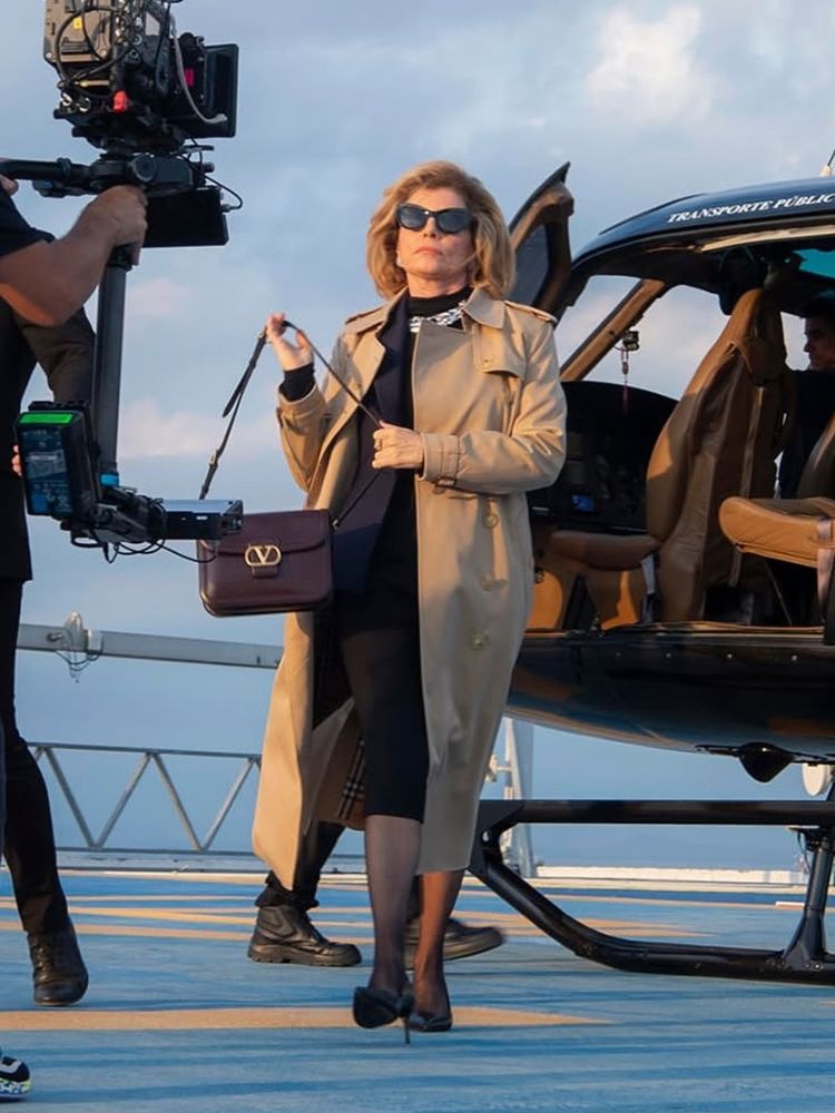 Débora Bloch ao lado de helicóptero usando vestido midi preto, joias e sobretudo bege 