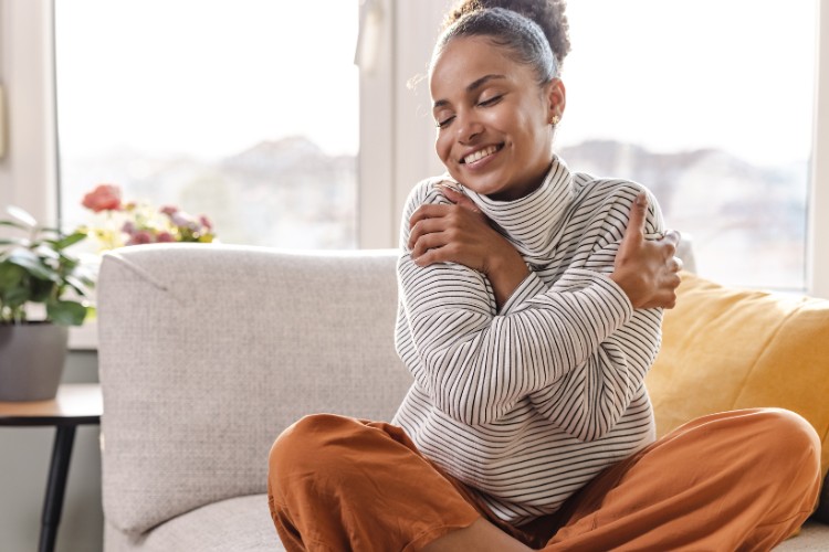 Mulher de pele negra no sofá, usando suéter, calça terracota, se abraçando e feliz