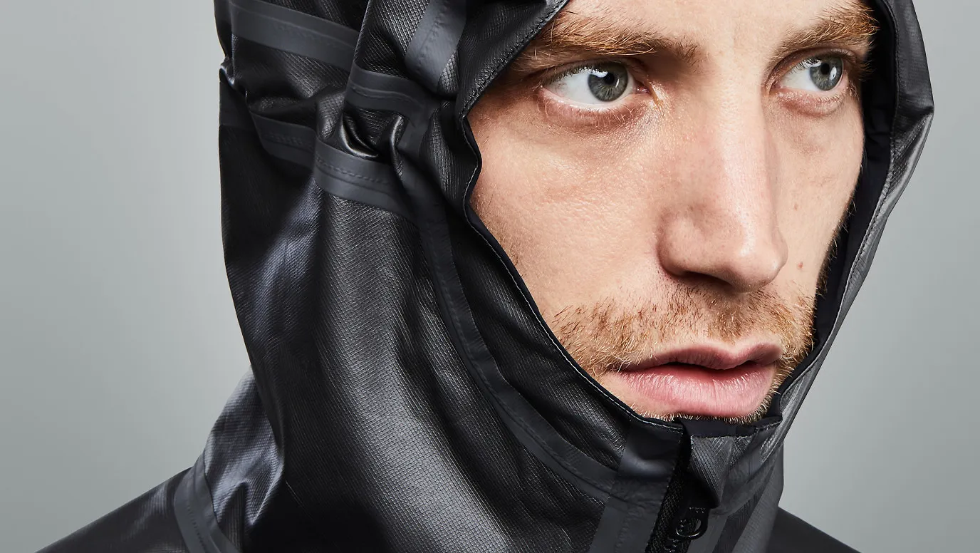 Close-up do rosto de um homem ruivo usando uma jaqueta de capuz cinza-escura. A jaqueta é feita de um material técnico e texturizado com costuras aparentes.