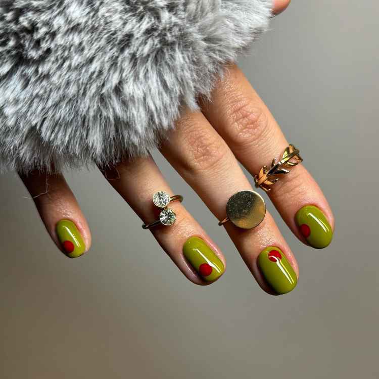 Unhas curtas pintadas de verde oliva com detalhes em vermelho. A mão está adornada com anéis dourados e prateados, incluindo um com design de folha. O fundo neutro e uma suave manga de pele cinza destacam o visual elegante e moderno.