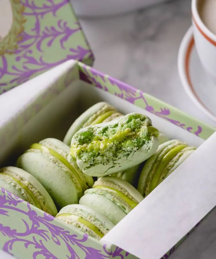 Uma caixa decorativa contém vários macarons verdes, com um macaron parcialmente mordido no topo, revelando seu recheio. A caixa está sobre uma superfície de mármore ao lado de uma xícara de café, criando uma cena elegante e apetitosa