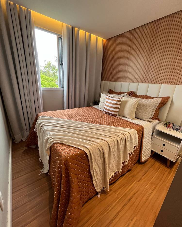A imagem mostra um quarto bem iluminado com uma cama grande. A cama é coberta por uma colcha marrom texturizada e adornada com vários travesseiros decorativos. A parede atrás da cama tem um design texturizado que complementa a cor e o padrão da colcha. Há cortinas cinza escuras penduradas em uma janela ao lado da cama, permitindo a entrada de luz natural. Um criado-mudo branco com gavetas está localizado ao lado da cama. O piso de madeira escura adiciona um toque de elegância ao quarto. O quarto é moderno e aconchegante, com iluminação suave.


