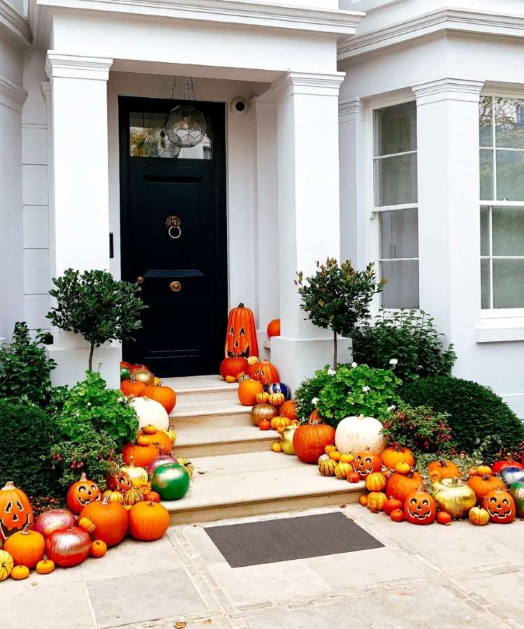 Entrada de casa com paredes brancas, porta preta, plantas e muitas abóboras na escada