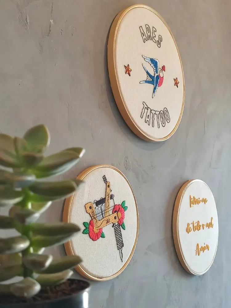 Três bordados em bastidores circulares pregados em uma parede cinza com uma planta próxima.