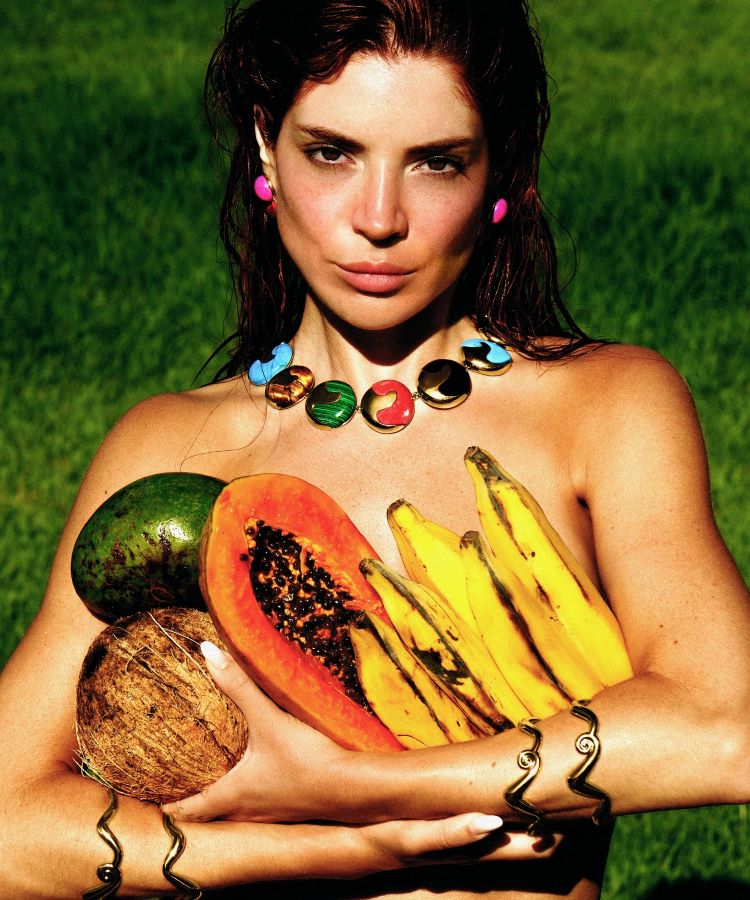 Mulher segurando frutas tropicais (mamão, banana, coco e abacate) em um gramado verde. Usa colar colorido, brincos rosa e pulseiras douradas, destacando um visual vibrante e natural em contraste com o fundo ensolarado.