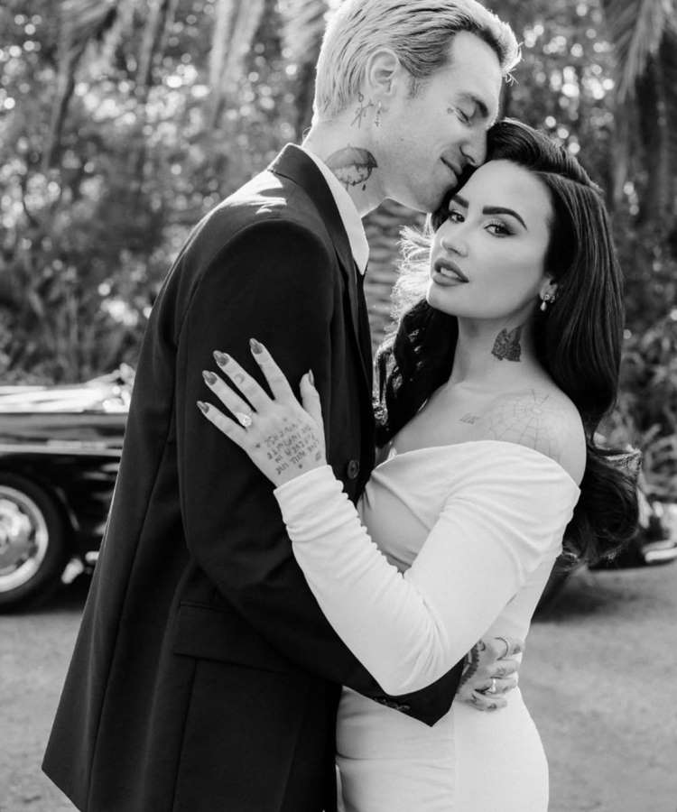 Em preto e branco, Demi Lovato e seu parceiro se abraçam. Ele inclina a cabeça para beijar a testa dela, enquanto ela olha para a câmera. Ambos estão próximos a um carro vintage em um ambiente externo com vegetação.