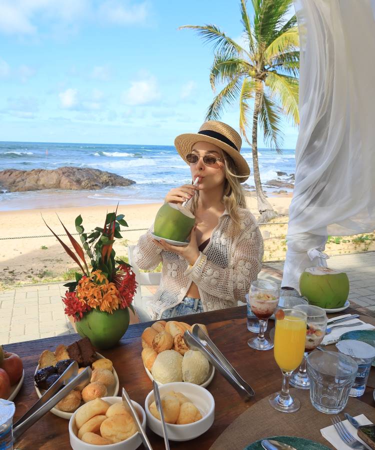 A imagem mostra Tata Estaniecki de chapéu de palha e óculos de sol, bebendo água de coco em uma mesa de café da manhã na praia. Ao fundo, coqueiros, areia e o mar com ondas. A mesa está repleta de pães, frutas e bebidas.