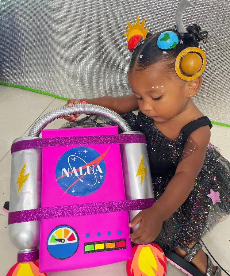 A imagem mostra uma criança pequena com uma mochila temática de astronauta. A mochila é rosa brilhante com detalhes prateados, simulando um traje espacial. Ela tem um logo parecido com o da NASA e elementos que lembram um painel de controle. A criança usa um penteado elaborado com enfeites coloridos que remetem a planetas e estrelas, combinando com o tema espacial da mochila.


