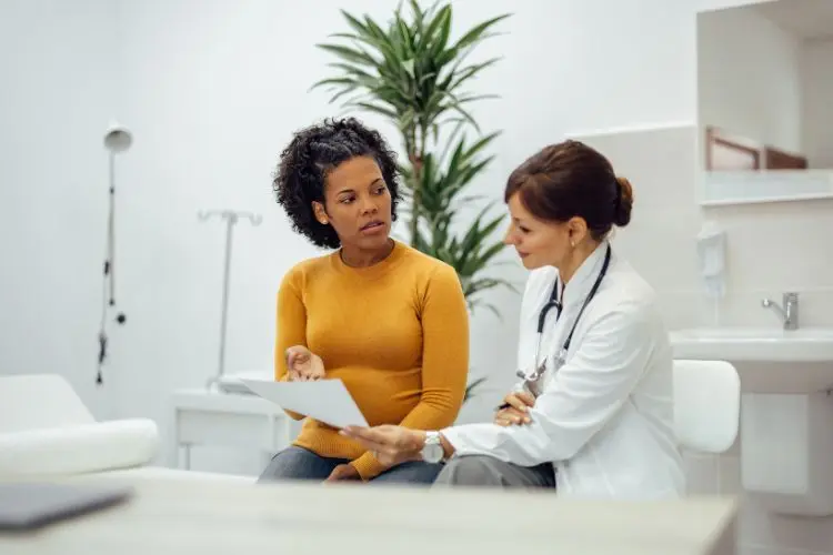 Mulher de pele negra usando blusa de manga comprida amarela sentada ao lado de médica de pele clara usando jaleco e segurando papéis