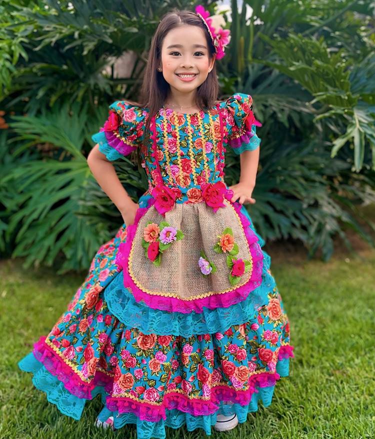 Menina usando traje junino rosa e azul, com flores, em fundo com planta costela de adão e chão gramado