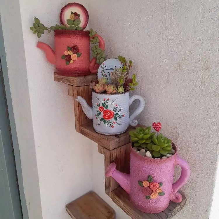 Três prateleiras em formato de escada com três vasos em formato de regador com suculentas dentro. 