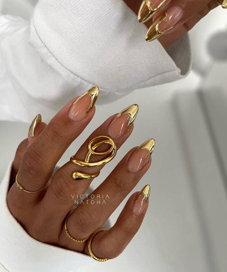 A imagem destaca unhas longas em formato stiletto, decoradas com pontas metálicas douradas sobre base nude translúcida. A mão exibe anéis dourados modernos e delicados, enquanto a manga branca realça o contraste elegante e sofisticado do design minimalista.