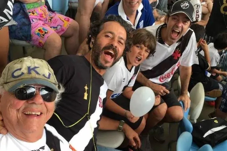 Três gerações de torcedores do Vasco da Gama, vestindo as camisas do time, sorriem e gritam na arquibancada. Um homem mais velho e de óculos escuros tira uma selfie com outros dois homens e uma criança. famosos que são parentes