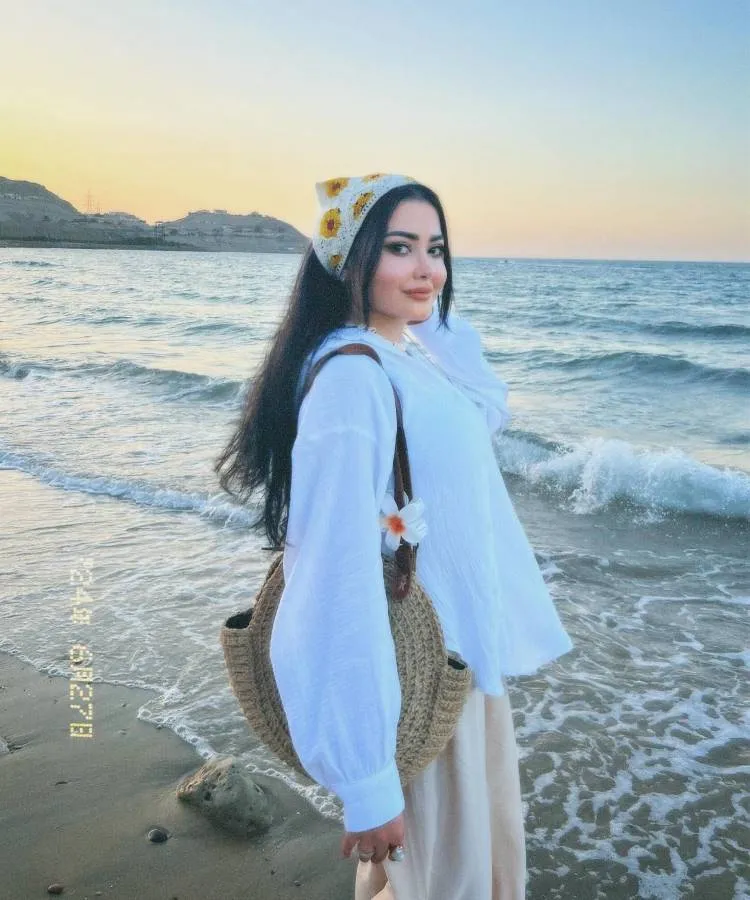 Uma mulher de cabelos longos e escuros, usando uma blusa branca e uma bandana amarela com flores, está na praia ao pôr do sol. Ela carrega uma bolsa de palha e sorri suavemente, com o mar e montanhas ao fundo.




