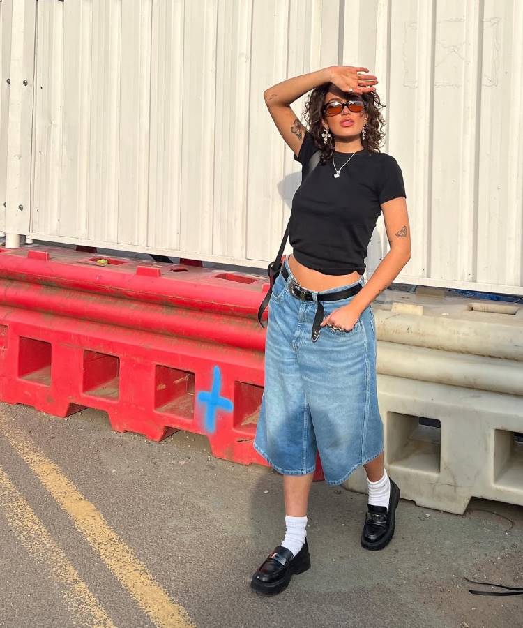 Uma mulher com cabelos cacheados, usando óculos de sol, camiseta preta, jorts, cinto preto, meias brancas e sapatos pretos. Ela cobre o rosto do sol e está ao lado de barreiras de construção vermelhas.