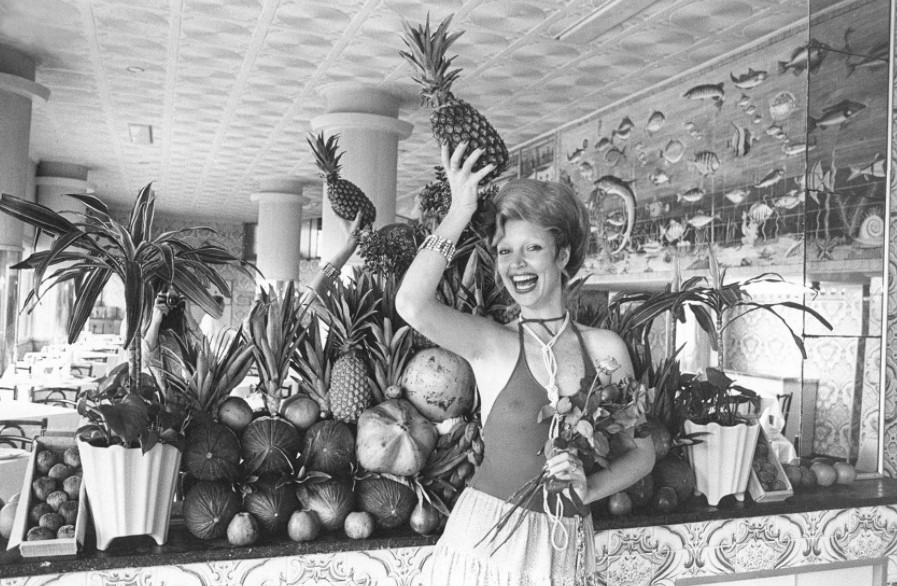 Foto em preto e branco de Elke Maravilha posando com um vestido decotado e segurando um abacaxi na cabeça diante de um espelho rodeado de frutas e plantas. Foto em preto e branco de Elke Maravilha posando com um vestido decotado e segurando um abacaxi na cabeça diante de um espelho rodeado de frutas e plantas.