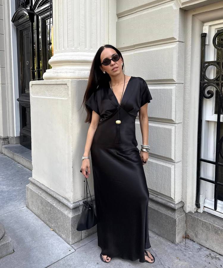 Uma mulher elegante com óculos de sol, vestindo um longo vestido de seda preta com decote em V e chinelos. Ela usa joias prateadas, segura uma pequena bolsa preta e está encostada em uma coluna de pedra.