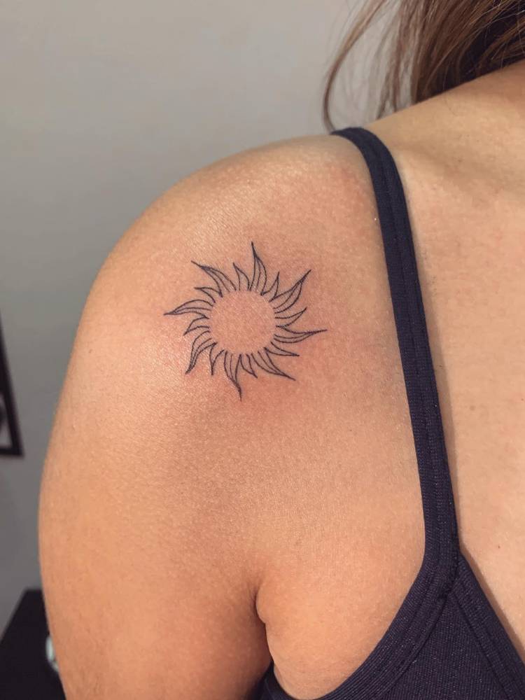 Mulher de pele clara usando regata preta e com tatuagem fine line no ombro, do sol estilo Enrolados