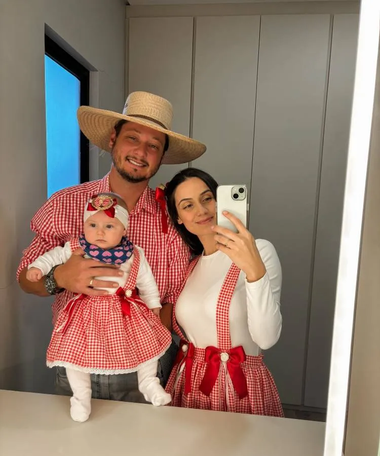 Família de três pessoas em trajes festivos. Homem com camisa xadrez e chapéu de palha, mulher com blusa branca e suspensórios xadrezes, e bebê com vestido xadrez e faixa na cabeça. Estão em frente a um espelho bem iluminado.