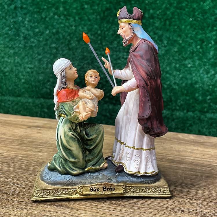 A imagem mostra São Brás abençoando uma criança, segurada por uma mulher ajoelhada. Ele veste trajes episcopais, com manto vermelho e mitra, enquanto segura velas cruzadas. A base dourada exibe o nome "São Brás", destacando a cena milagrosa.