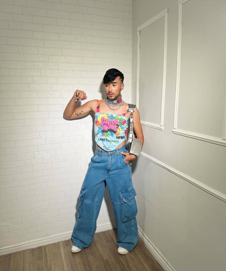 A imagem mostra um homem estiloso usando um abadá customizado com cores vibrantes, incluindo rosa, azul e verde, decorado com estampas tropicais e frases. Ele combina o look com calça jeans larga, acessórios prateados e uma bolsa metálica.