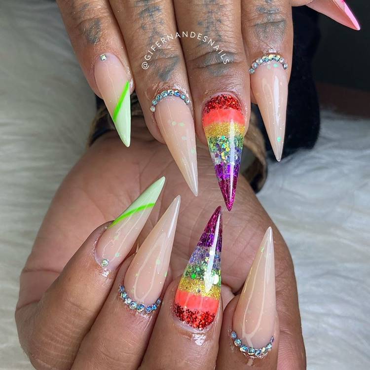 Mão com unhas decoradas coloridas: Polegar: nude com rosa e pedrinha na cutícula Indicador:nude com pedrinha em toda cutícula Médio: arco-íris de glitter Anelar: nude com pedrinha em toda cutícula Mínimo nude com verde e pedrinha na cutícula