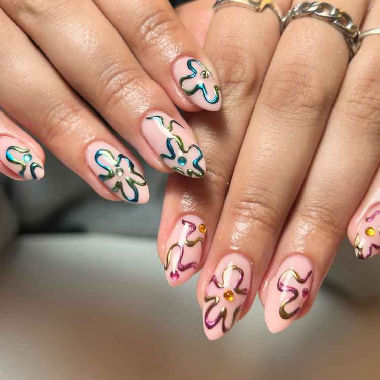 Unhas longas decoradas com esmalte nude e desenhos florais estilizados, contornados em azul, verde e rosa metálico. Pedras brilhantes adornam o centro de cada flor. As mãos, com anéis prateados, estão entrelaçadas sobre um fundo neutro, destacando o design criativo.