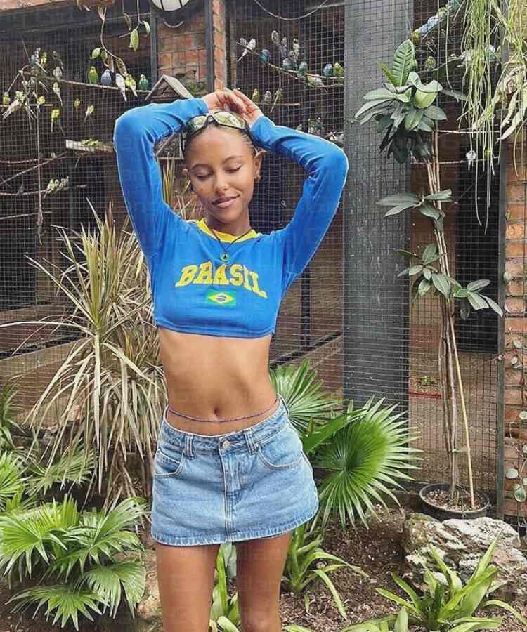 Imagem com fundo verde com pássaros presos em grade. No centro, mulher usando cropped de manga longa azul do Brasil + mini saia