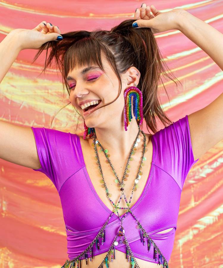 A mulher sorri alegremente, usando um top roxo vibrante e brincos coloridos de miçangas em forma de arco-íris. Seu cabelo está preso em rabos de cavalo altos, complementado por maquiagem rosa brilhante, em um cenário carnavalesco rosa e dourado.