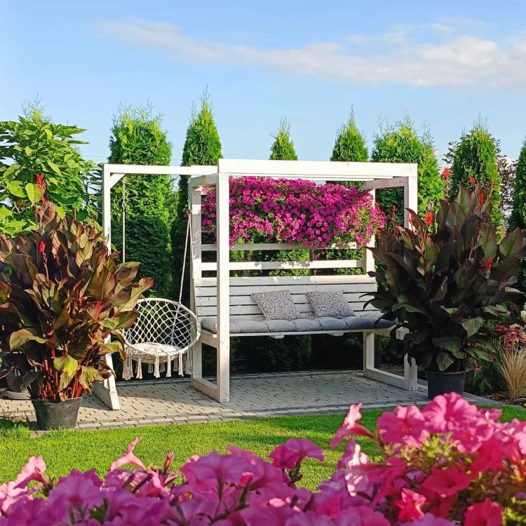A imagem exibe um jardim encantador com um banco de madeira branco decorado com almofadas cinzas. Acima, flores rosa vibrantes adornam a estrutura. Plantas altas e arbustos cercam o cenário, enquanto um balanço de macramê complementa o ambiente relaxante.