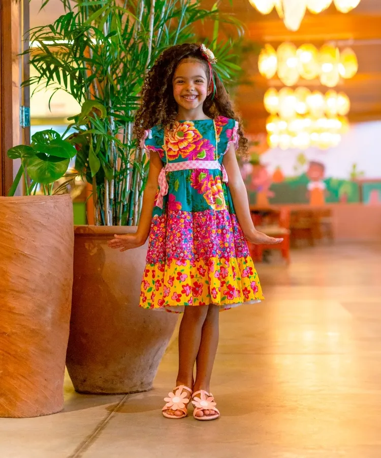 Uma menina sorridente, com cabelos cacheados castanhos e pele morena clara, posa em um ambiente interno decorado com plantas e iluminação suave. Ela veste um vestido colorido com estampas florais em tons de azul, rosa e amarelo, adornado com uma faixa rosa na cintura e detalhes florais nos ombros. Sandálias rosa completam o visual alegre e infantil. A atmosfera parece acolhedora e festiva.

