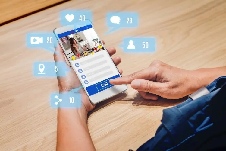 A imagem mostra uma pessoa segurando um smartphone exibindo uma transmissão ao vivo. Na tela, há interações sociais: curtidas, comentários e visualizações. Ícones flutuantes indicam 43 curtidas, 23 comentários, 50 espectadores, e outros ícones sociais. O ambiente é uma mesa de madeira.